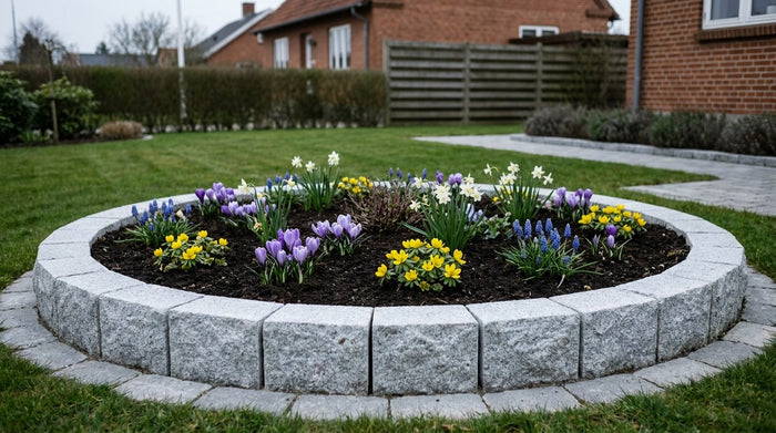 Et færdigbygget rundt højbed i lys grå portugisisk granit med frodige grønne planter i en dansk have
