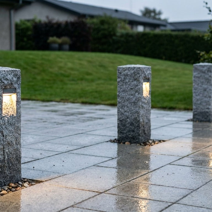 En smukt oplyst dansk indkørsel med lyse granitskærver og solide mørke granitpullerter, der markerer overgangen til græsplænen i mørket.