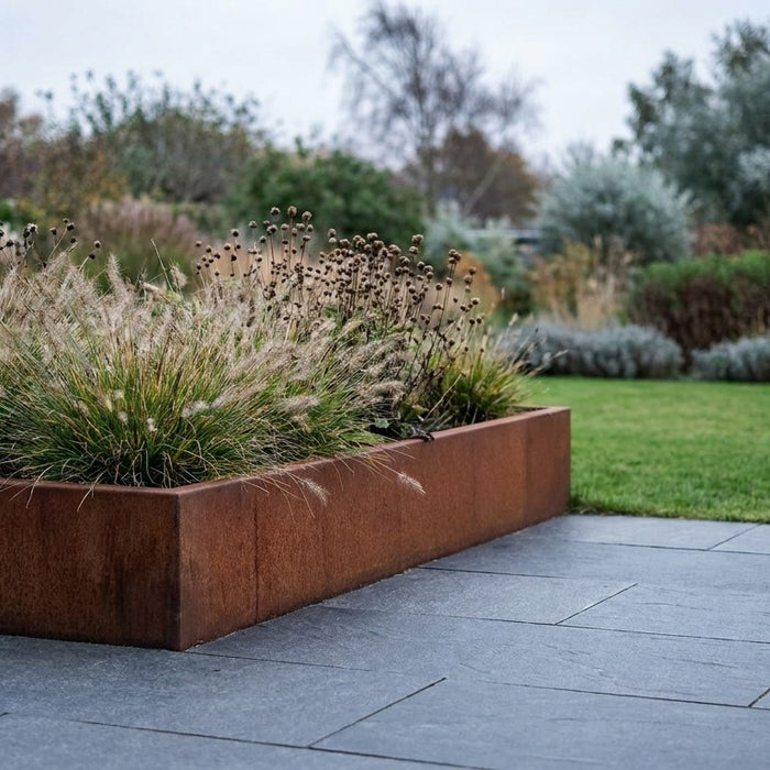 Et smukt cortenstål højbed fyldt med frodige grønne planter, der skaber kontrast til en terrasse af grå natursten