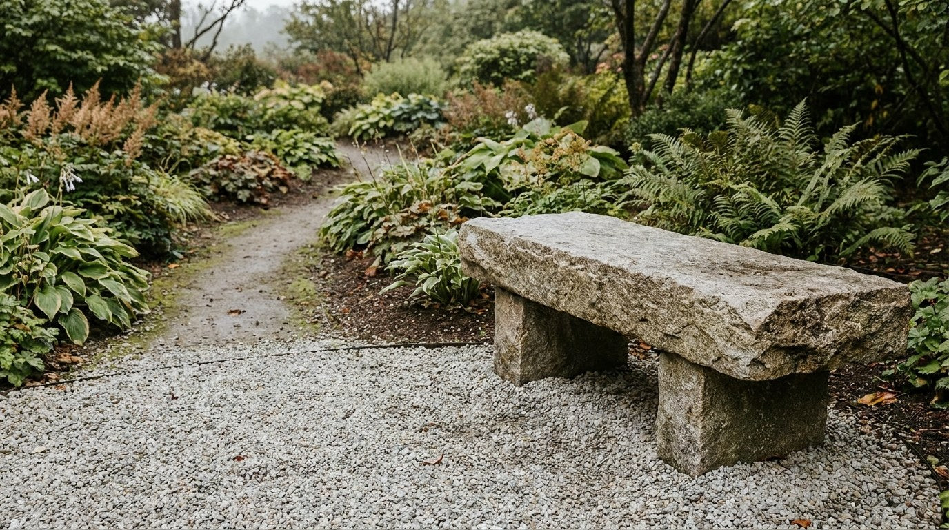 En massiv og elegant granitbænk placeret på et stabilt underlag af chaussesten for enden af en havesti omgivet af grønne planter