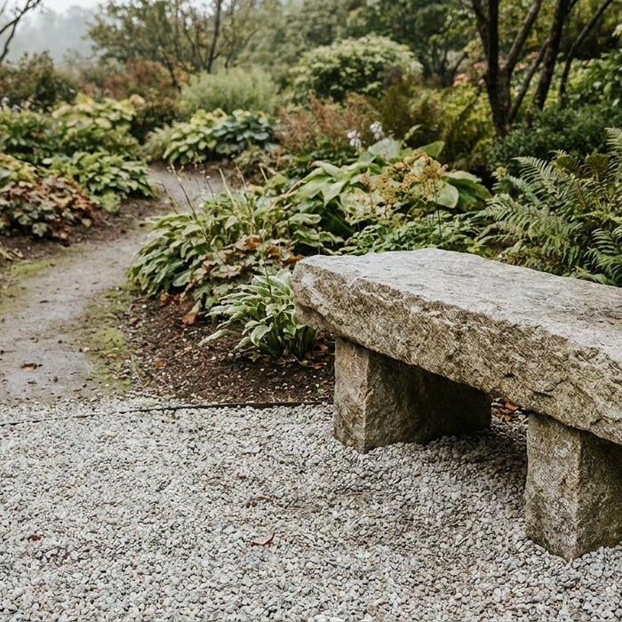 En massiv og elegant granitbænk placeret på et stabilt underlag af chaussesten for enden af en havesti omgivet af grønne planter