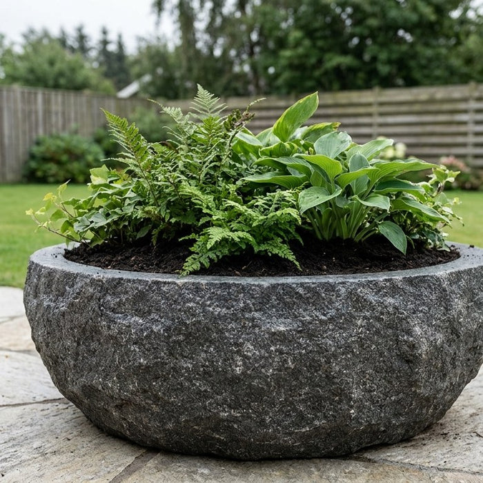 Nærbillede af hænder der fylder luftig og næringsrig plantejord i en stor udendørs plantekumme af beton over et drænlag af granitskærver.