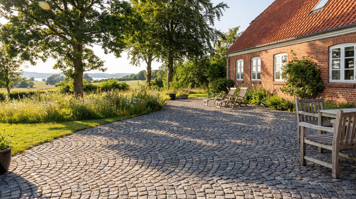 Chaussestensbelægning foran rødstenshus med have og udsigt.